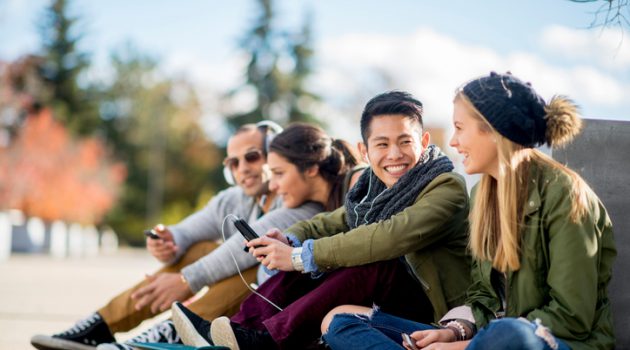 A multi-ethnic group of college age students are sitting together texting on their cell phones and taking selfies together.